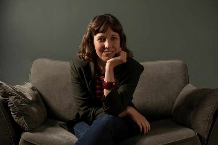 A portrait of a White woman sitting on a couch, smiling, with one hand resting under her chin and wearing a dark blazer.