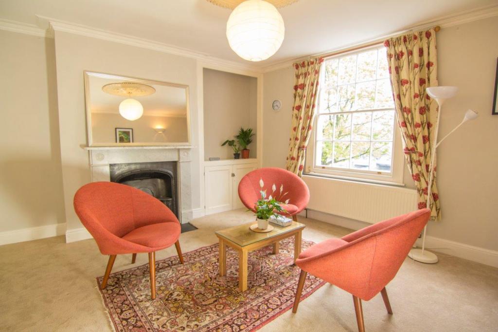 A bright and inviting therapy room featuring two red chairs, a small wooden table with a potted plant, and soft lighting from a ceiling fixture. Large windows allow natural light to enter, creating a calm and welcoming atmosphere.