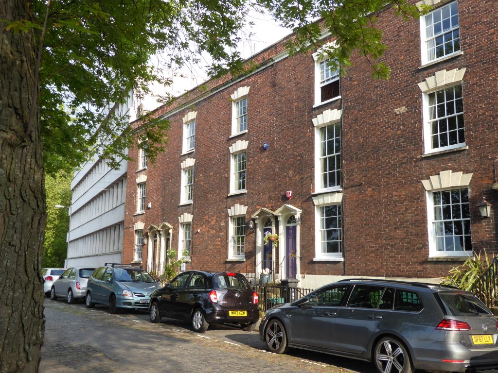 A street view of a brick building with large windows and a colorful door, flanked by parked cars along a cobblestone road, and surrounded by trees.